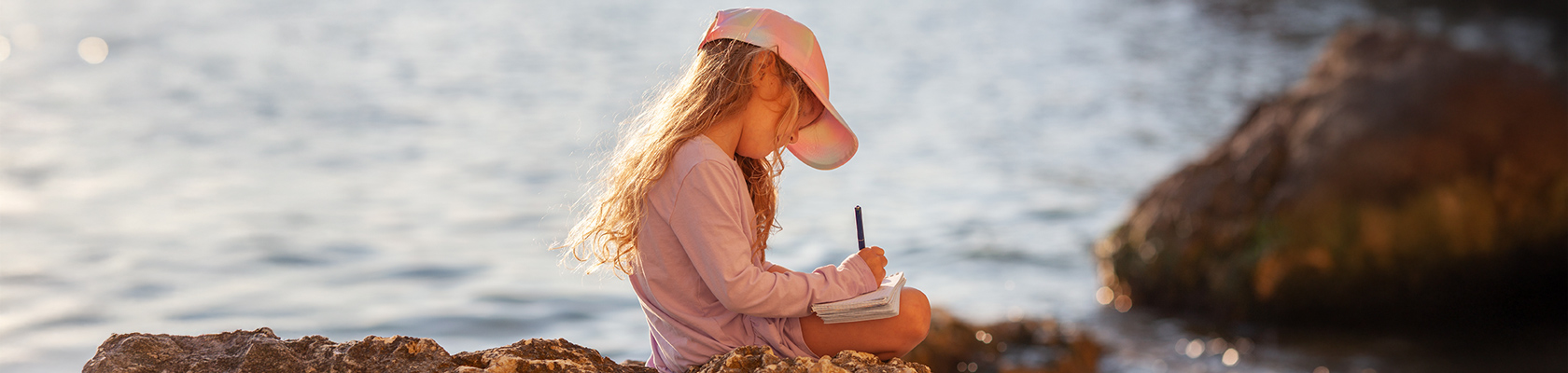 Une jeune petite fille blonde en train de noter des choses sur son carnet de voyage Moby Lines sur une plage Corse, assise sur des rochers au pied de l’eau.